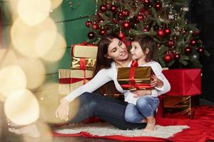 Holiday is unite people. Cheerful mother and daughter sitting near the Christmas tree that behind. Cute portrait photo