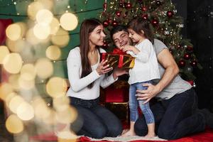 compartiendo vacaciones con hija. Encantadora familia se sienta cerca del árbol de Navidad con cajas de regalo en la noche de invierno, disfrutando del tiempo que pasan juntos