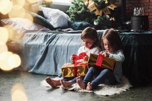 tira de esas cosas rojas para abrir. vacaciones navideñas con regalos para estos dos niños que se sientan en el interior de la bonita habitación cerca de la cama foto