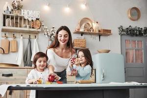Young beautiful woman feeds two kids with apples while they sitting near the table with toys photo
