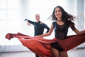 Girl is spin around herself. Young pretty woman in red and black clothes dancing with bald guy in the white room