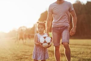 preguntándose hasta dónde esta chica puede patear esa pelota. Foto de papá con su hija en la hermosa hierba y bosques al fondo