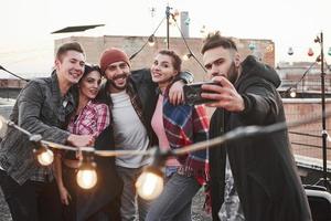 Grupo de jóvenes amigos alegres que se divierten, se abrazan y se toman selfie en el techo con bombillas decorativas
