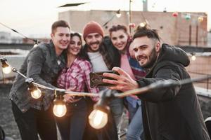 Focus on the smartphone. Group of young cheerful friends having fun, hug each other and takes selfie on the roof with decorate light bulbs