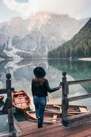 se sostiene en un muelle de madera. Mujer con sombrero negro disfrutando del majestuoso paisaje de montaña cerca del lago con barcos