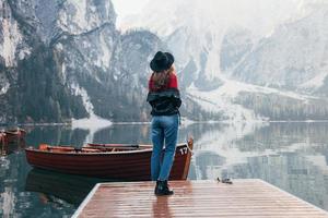 foto desde atrás. Mujer con sombrero negro disfrutando del majestuoso paisaje de montaña cerca del lago con barcos
