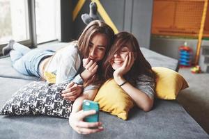 dos lindas hermanas gemelas sonrientes sosteniendo smartphone y haciendo selfie. las niñas se acuestan en el sofá posando y alegría foto