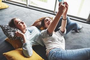 Two cute smiling twins sisters holding smartphone and making selfie. Girls lie on the couch posing and joy