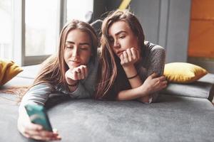 Two cute smiling twins sisters holding smartphone and making selfie. Girls lie on the couch posing and joy photo