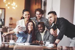 Friends having fun at restaurant. Three boys and two girls making selfie and laughing. On foreground boy holding smart phone. All wear casual clothes photo