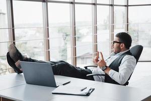 Beautiful young businessman with glasses holding his legs on the table looking at a laptop in the office photo