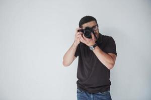 A young hipster man in eyepieces holds a DSLR camera in hands standing against a white wall background photo