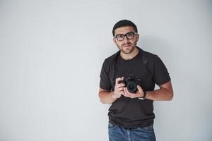 A young hipster man in eyepieces holds a DSLR camera in hands standing against a white wall background photo