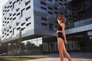 sexy mujer joven con gafas de sol y traje de baño negro está de pie en el fondo de los edificios de la ciudad durante la puesta de sol foto