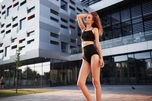 Sexy young woman in sunglasses and black bathing suit is standing on the city buildings background during sunset