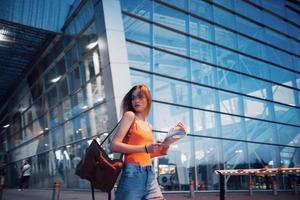 A young beautiful girl with a backpack behind her shoulders standing on the street near an airport. She just arrived from the rest and very happy photo