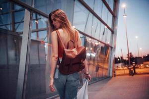 A young beautiful girl with a backpack behind her shoulders standing on the street near an airport. She just arrived from the rest and very happy photo