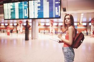 Hermosa joven turista con mochila en el aeropuerto internacional, cerca del tablero de información de vuelos foto