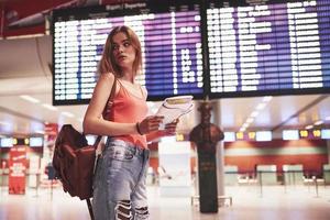 Beautiful young tourist girl with backpack in international airport, near flight information board photo