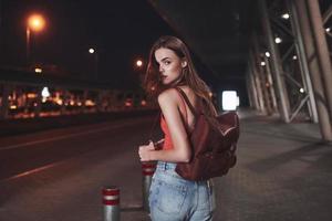 A young beautiful girl with a backpack behind her shoulders stands on the street near an airport or a railway station on a warm summer evening. She just arrived and waits for a taxi or her friends photo