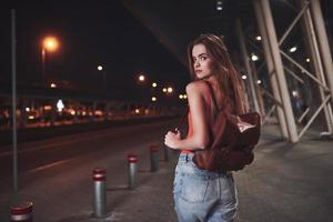 A young beautiful girl with a backpack behind her shoulders stands on the street near an airport or a railway station on a warm summer evening. She just arrived and waits for a taxi or her friends photo