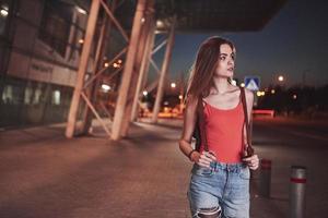 A young beautiful girl with a backpack behind her shoulders stands on the street near an airport or a railway station on a warm summer evening. She just arrived and waits for a taxi or her friends photo