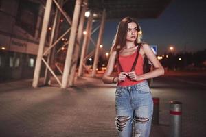 A young beautiful girl with a backpack behind her shoulders stands on the street near an airport or a railway station on a warm summer evening. She just arrived and waits for a taxi or her friends photo