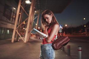 Young girl costs at night near the terminal of the airport or station and reading city map and looking for hotel. Cute tourist with backpacks determine the concept of travel