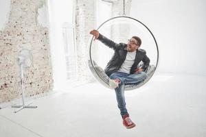 Full-length portrait of relaxed hipster man in casual sitting on hanging chair bullet and smiling on camera isolated over white background photo