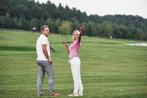 Woman showing something behind. Couple of golf players with sticks in their hands standing on the lawn