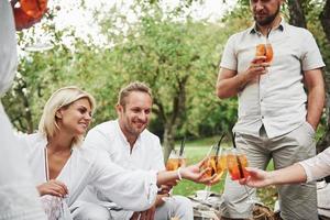 Inside of the cheerful atmosphere. Friends drinks brand new fresh cocktails when sit on the field with trees photo