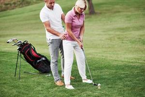 Now try on your own. Man teaching woman to play golf at the sport field with equipment behind