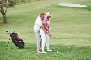 Man teaching woman to play golf at the sport field with equipment behind photo