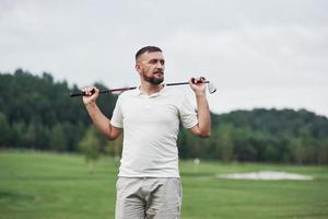 Looking far away. Portrait of standing golf player in the lawn and stick in hand. Woods at background