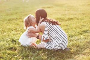 Mother and daughter sitting on the grass of the field and kiss each other