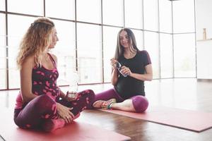 Female pregnant friends having conversation while sitting in the hall on fitness mat and holding bottles of water photo