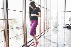 Morena embarazada de pie en el gimnasio cerca de las ventanas y manteniendo las manos en el vientre foto