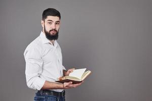 Portrait of a bearded young man wearing a white shirt and holding an open planner and a pen. A gray wall background photo