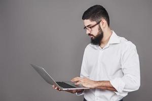 Handsome business man in glasses and suit holding laptop in hands and writing something. Side view. Isolated gray background photo