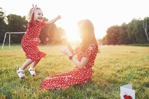 Jumping with hands raised up. Girl with her daughter having fun with the bubbles outside sitting on the grass