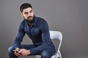 Portrait of toothy handsome bearded man sitting on a chair with phone on hands photo