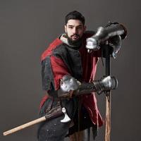 Medieval knight on grey background. Portrait of brutal dirty face warrior with chain mail armour red and black clothes and battle axe photo