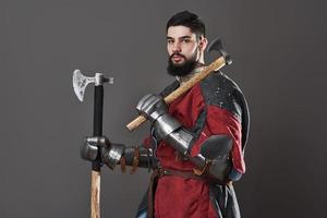 Medieval knight on grey background. Portrait of brutal dirty face warrior with chain mail armour red and black clothes and battle axe photo