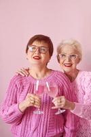 Dos mujeres mayores con estilo en suéteres de color rosa bebiendo vino rosado en la cocina moderna chismeando. amistad, hablar, chismes, eventos, relaciones, noticias, concepto de familia foto