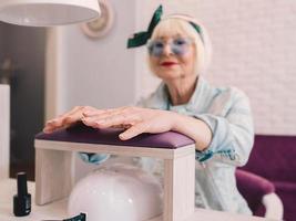 Maestro de manicura en guantes azules batiendo las manos de una anciana elegante con gafas de sol azules y chaqueta vaquera sentado en el salón de manicura foto