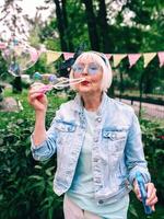senior stylish woman with gray hair and in blue glasses and jeans jacket blowing bubbles outdoors. Holidays, party, anti age, fun concept photo