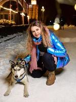 Happy smiling young Caucasian woman in scarf, hat, jacket, mittens with husky dog at winter night outdoor. New year, fun, friendship, winter concept