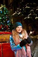 Happy smiling young Caucasian woman in scarf, hat, jacket, mittens with sparkler by the christmas tree outdoor. New year, fun, winter concept photo