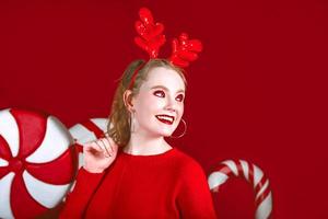 portrait of beautiful redhead funny christmas woman in deer horns with candy on the red background photo