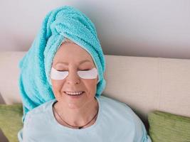 senior caucasian stylish woman laying on a coach with blue towel on her had and collagen eye patches. Anti age, healthy life style, photo